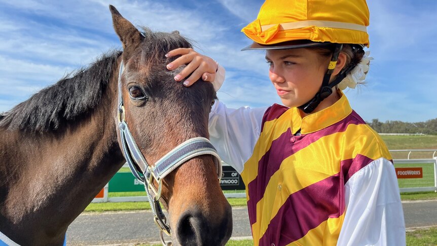 A girl wearing a brown and yellow jockey outfit pats a small brown horse