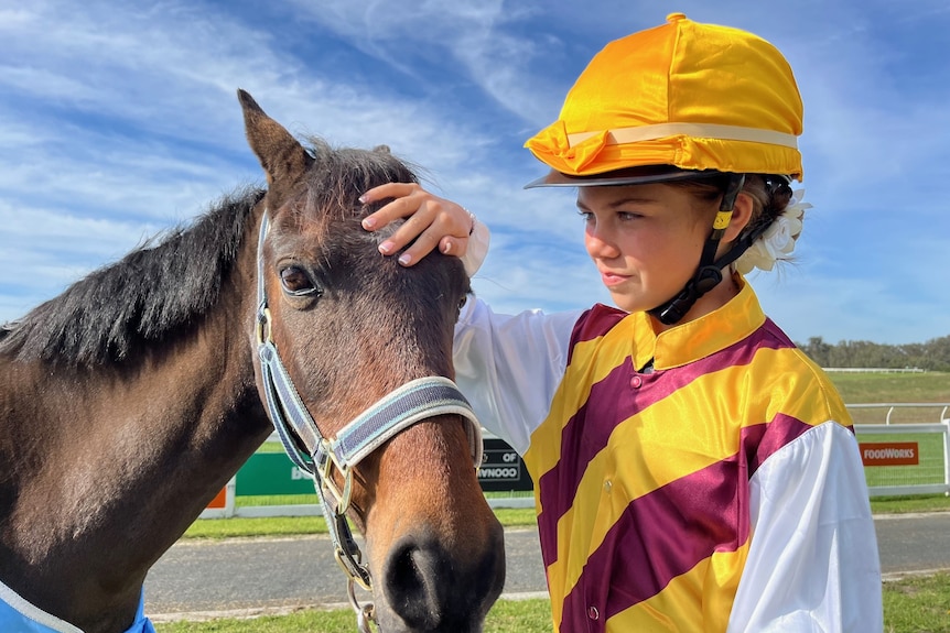 A girl wearing a brown and yellow jockey outfit pats a small brown horse