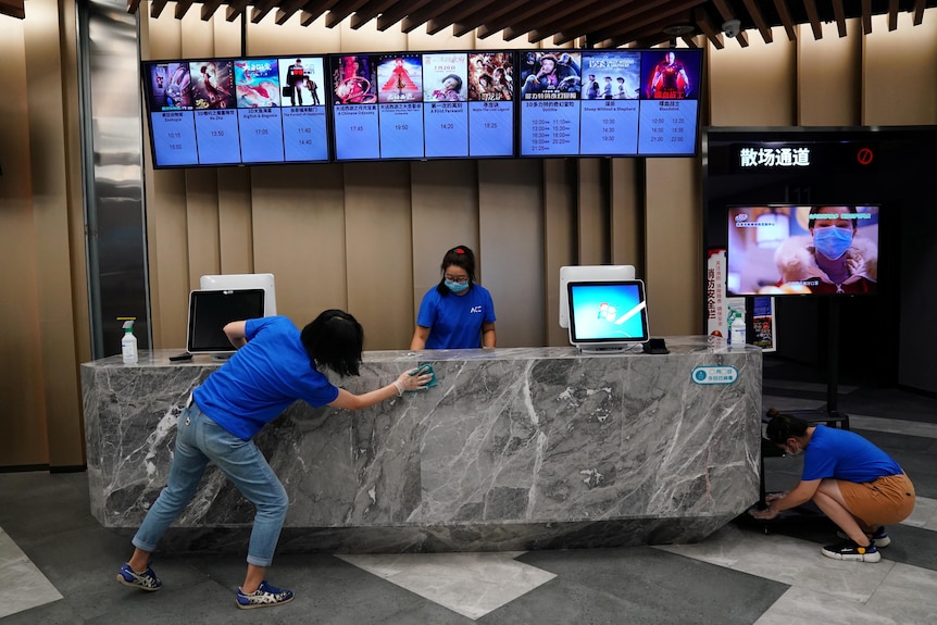 Three Chinese staff members clean the ticket desk at a cinema, with screens above that show film times