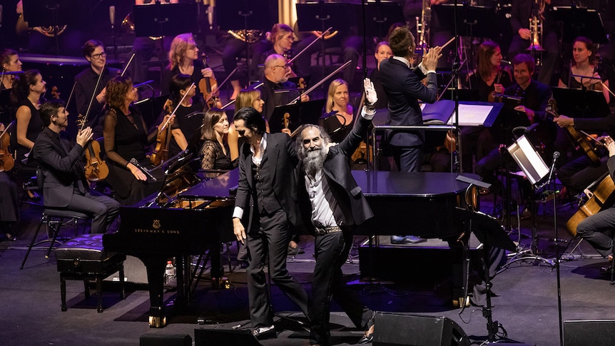 Nick Cave and Warren Ellis wave to the crowd, arm in arm, in front of an orchestra at the Sydney Opera House