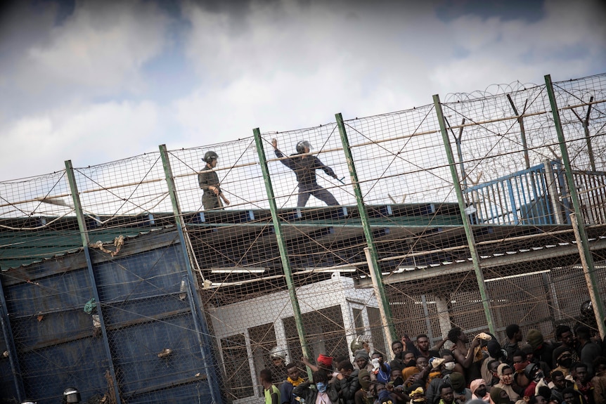 Riot police officers stand on a roof behind a crowd pushing through a fence
