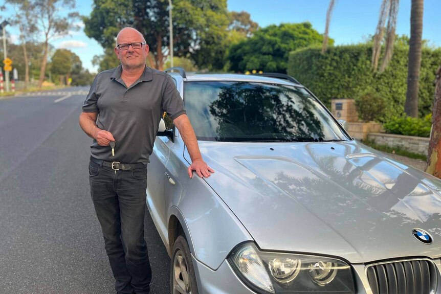A man in a grey shirt and black pants leans on a silver Mercedes car on a wide, leafy street.
