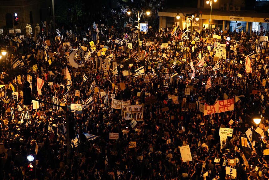 A large night-time crowd of protesters waving flags and signs outside a brick residence.