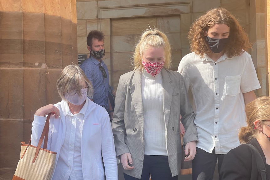 A blonde woman flanked by an older woman and a man with long hair outside a court building