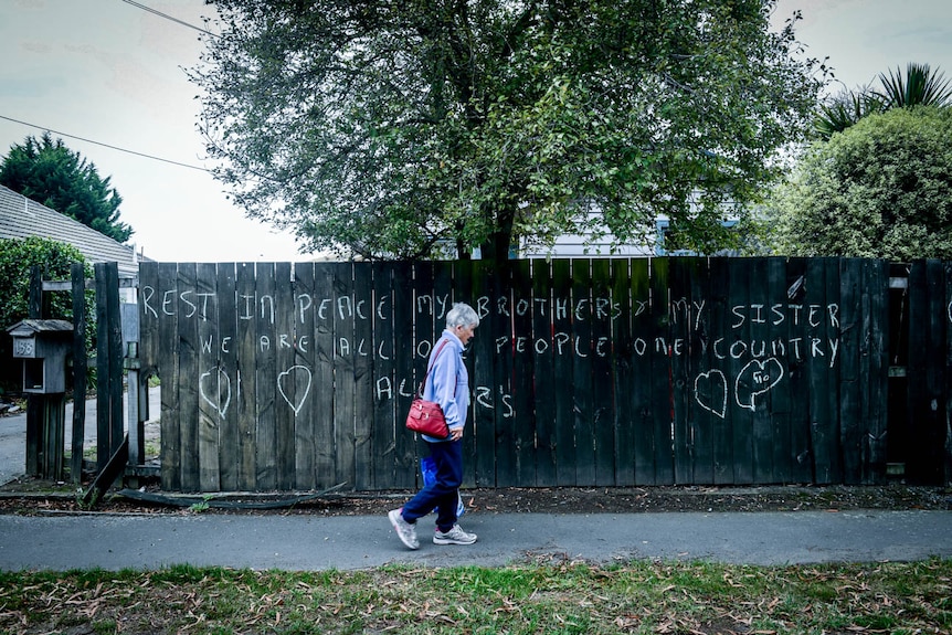 A woman walks past a fence which reads "rest in peace my brothers and sisters, we are all one people"