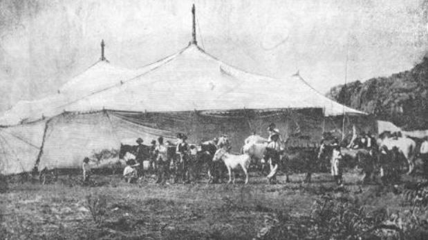 Old black-and-white photo of people and horses lined up outside a big white circus tent