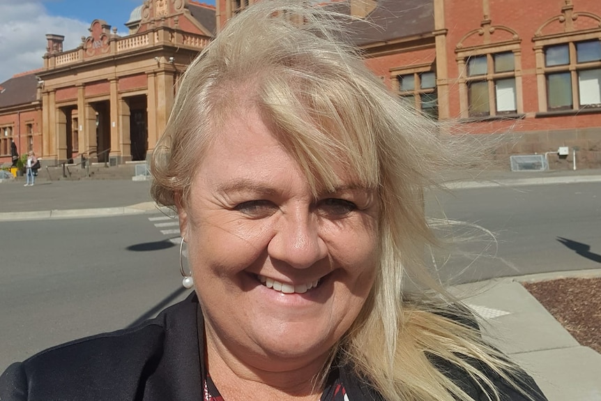 A woman with blonde hair takes a selfie in front of a building.