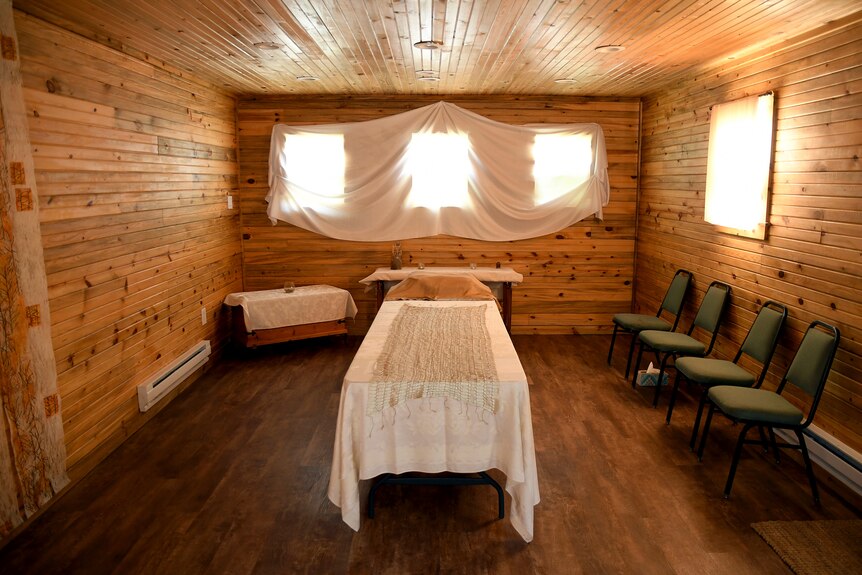 A bed in the middle of a wooden cabin room with sun shining through soft draped curtains 