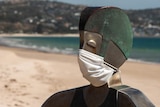 A metal sculpture of a lifeguard wearing a face mask in front of an empty beach