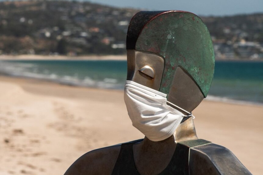A metal sculpture of a lifeguard wearing a face mask in front of an empty beach