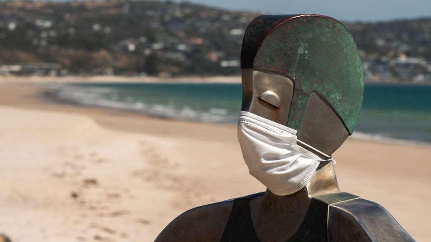 A metal sculpture of a lifeguard wearing a face mask in front of an empty beach