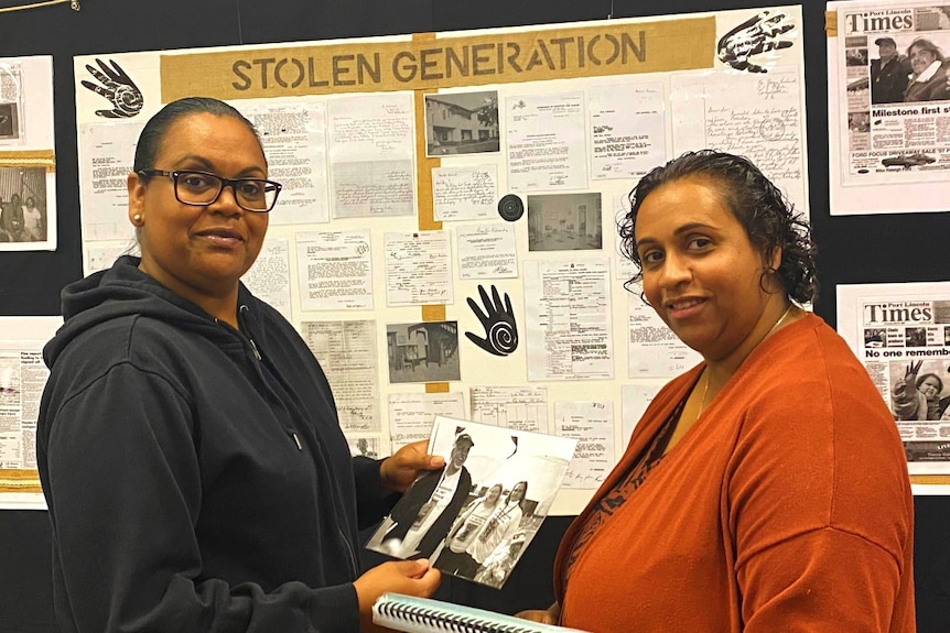 Two indigenous women holding a folder and photograph in front of display board for Stolen Generation