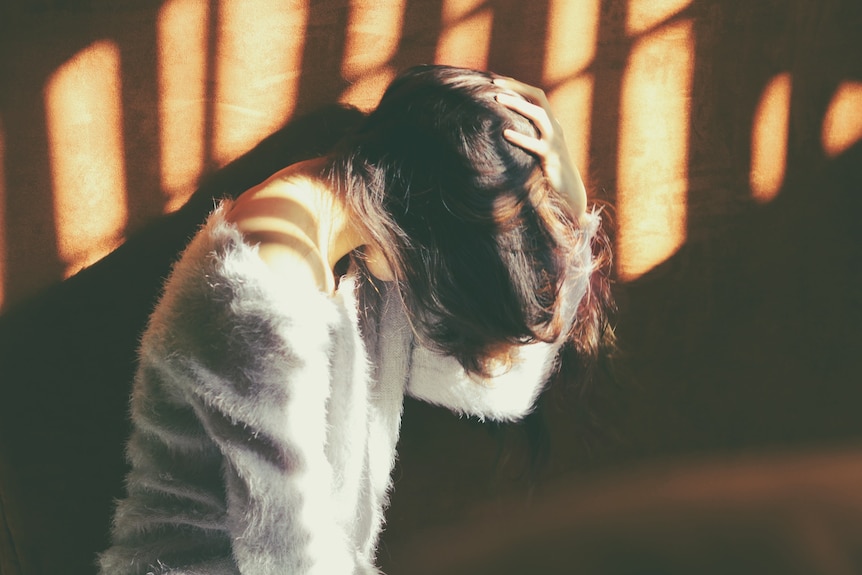 A woman with short dark hair who is wearing a grey sweater stands in a patch of sunlight with her hand to her head