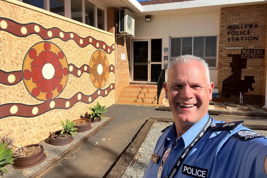 Superintendent Post out front of Mullewa police station which has an Indigenous mural on wall and plants underneath.