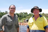 A man in high vis and a hat and a man in a khaki shirt stand smiling side by side in a field.