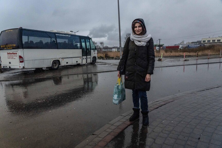 A woman wearing a parka, scarf and beanie stands on a road nearby a bus on a wet, grey day