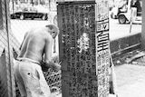 A black and white photo of an old man painting black calligraphy onto an electricity box on the street.