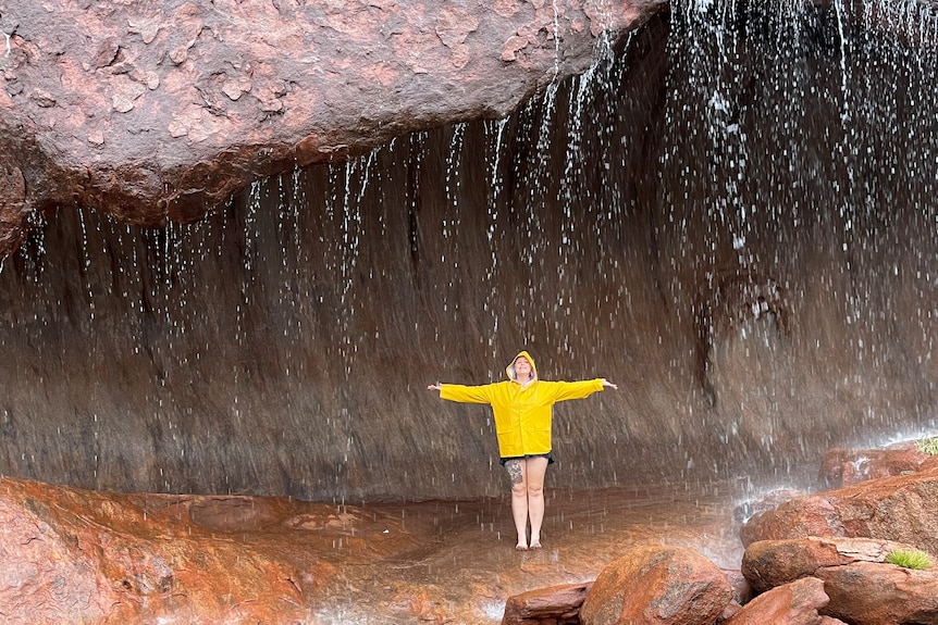 A woman in a bright yellow hooded raincoat stands with her arms out under a rock with water falling over it