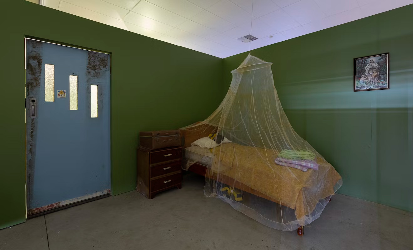 An installation artwork featuring a single bed, bedside table, and mosquito net in a dingy-looking dark room.