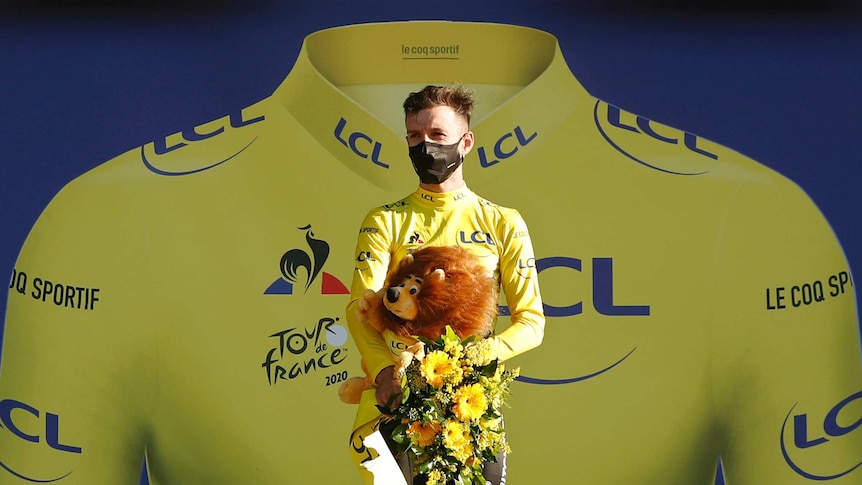 A cyclist stands on the podium in front of a large image of the yellow jersey at the Tour de France.