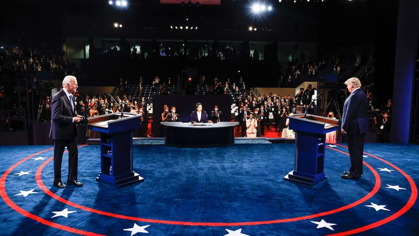 Joe Biden and Donald Trump stand far apart on a blue stage in front of a masked audience.