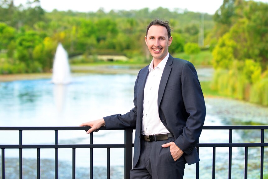 A man wearing a dark suit stands in front of a lake, smiling.