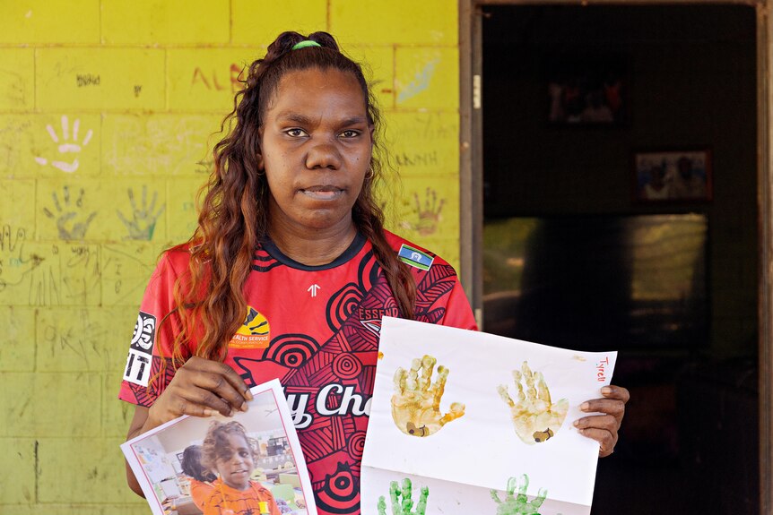 A young Aboriginal woman holds up a photo of a young child and painted handprints on paper