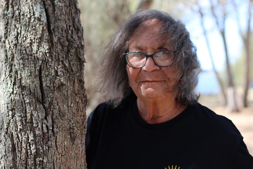 Elder Vivienne Mason stands next to a tree in a black shirt and glasses, her grey hair windswept across her face