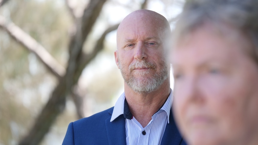 Bald man with white beard, blue collared shirt and navy jacket, looking at blurred out woman in the foreground