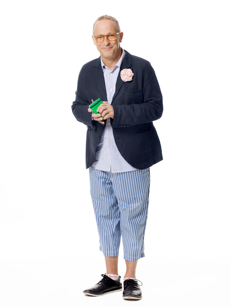 David Sedaris, dressed in a blazer and cropped, pinstripe pants, holds a notepad.