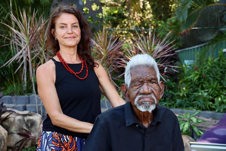 Woman wearing black top and red necklace stands behind indigenous man sitting on wheelchair