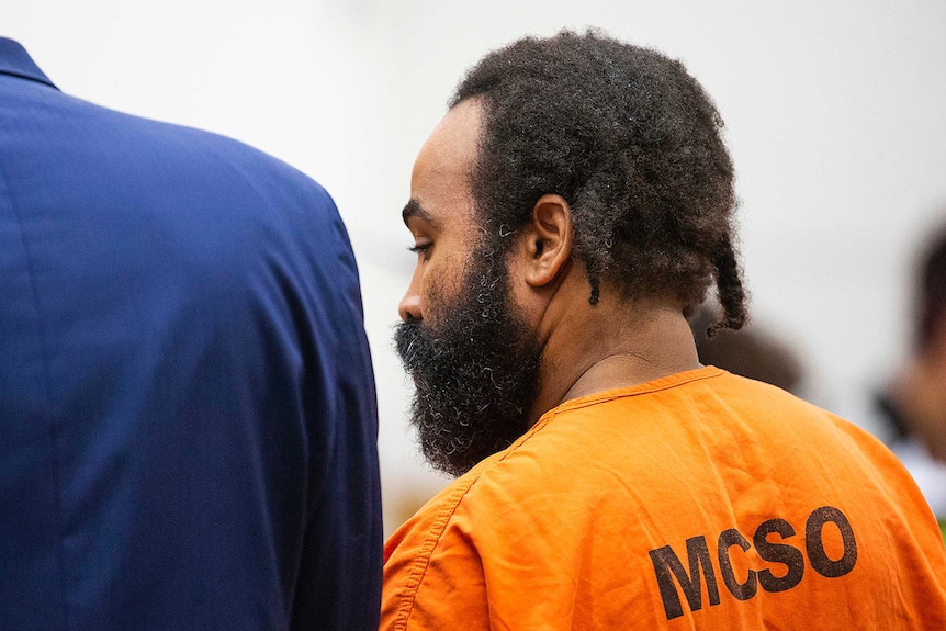 Nathan Sutherland stands in court facing away from the camera, wearing an orange prison jumpsuit.