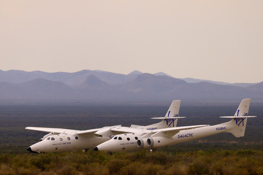 A white transport jet is flying close to the ground with a mountain range in the background.