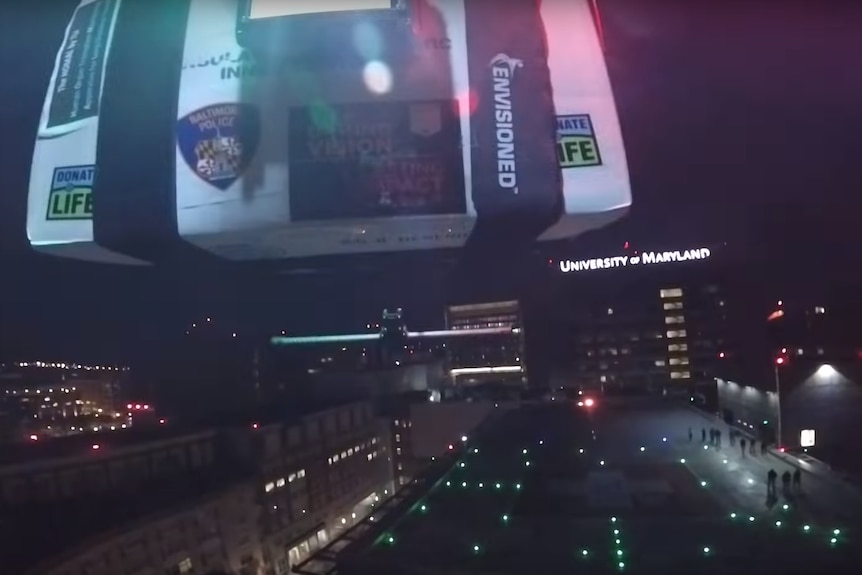 Vision showing a drone carrying a large white esky approaching a landing pad in the night sky.