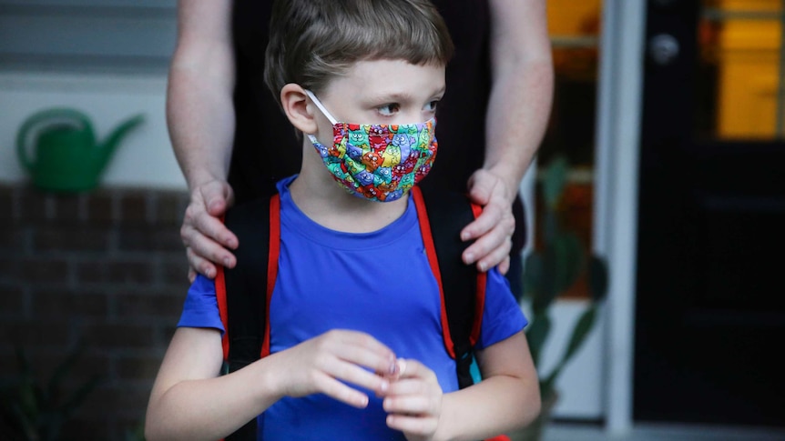A boy in a blue shirt and a backpack wears a bright, multi-coloured face mask.