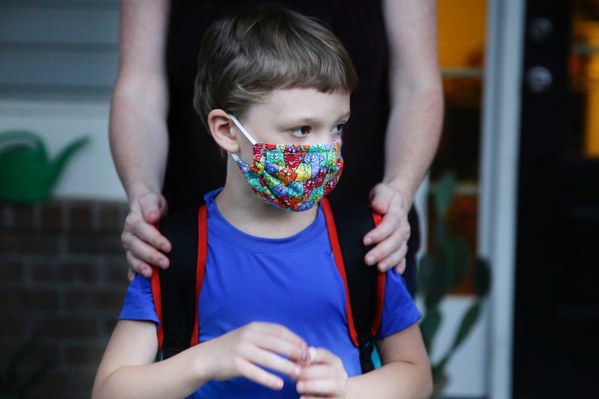 A boy in a blue shirt and a backpack wears a bright, multi-coloured face mask.