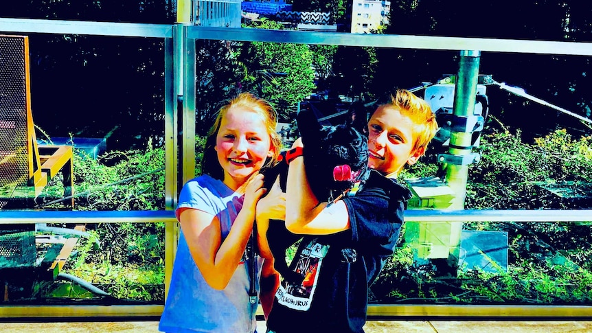 Two children cuddle a black dog with city skyscrapers in the background