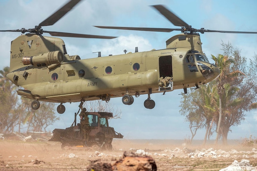 Soldiers connect equipment to a helicopter that hovers above the ground on an island