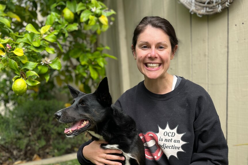 a woman sits on the grass in a backyard smiling and hugging a black dog, wearing a jumper that syas 'ocd is not an adjective'