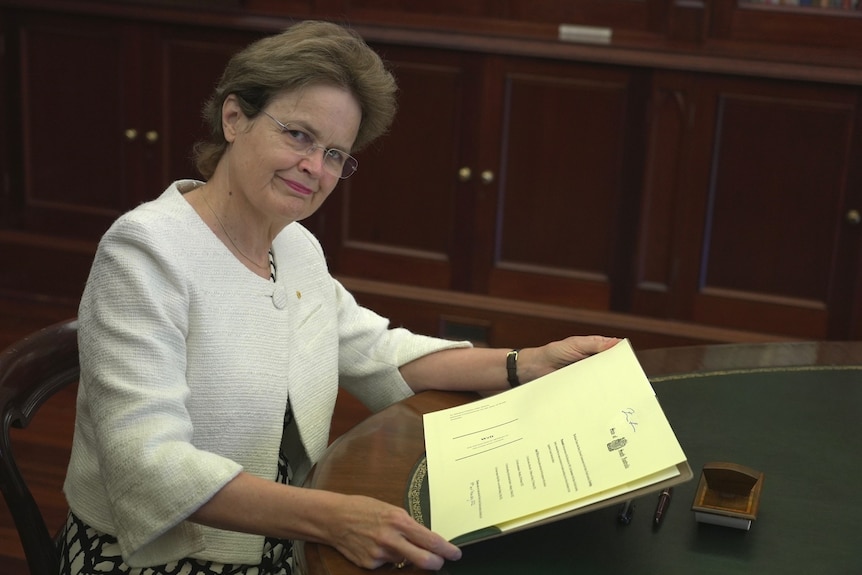 A woman wearing a white jacket sits at an oval desk holding a document