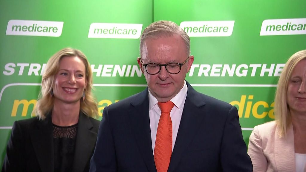 Anthony Albanese speaking in orange tie in front of two women and green medicare background.
