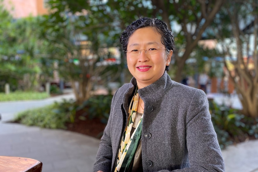 A young Asia woman with short hair wearing a grey coat sits outside with trees behind her. She is smiling at the camera.