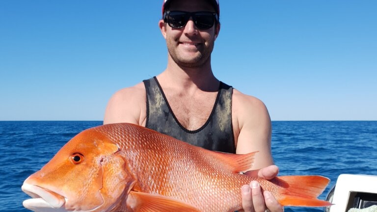 A smiling young man wearing a cap and sunglasses stands on a boat holding a large fish.