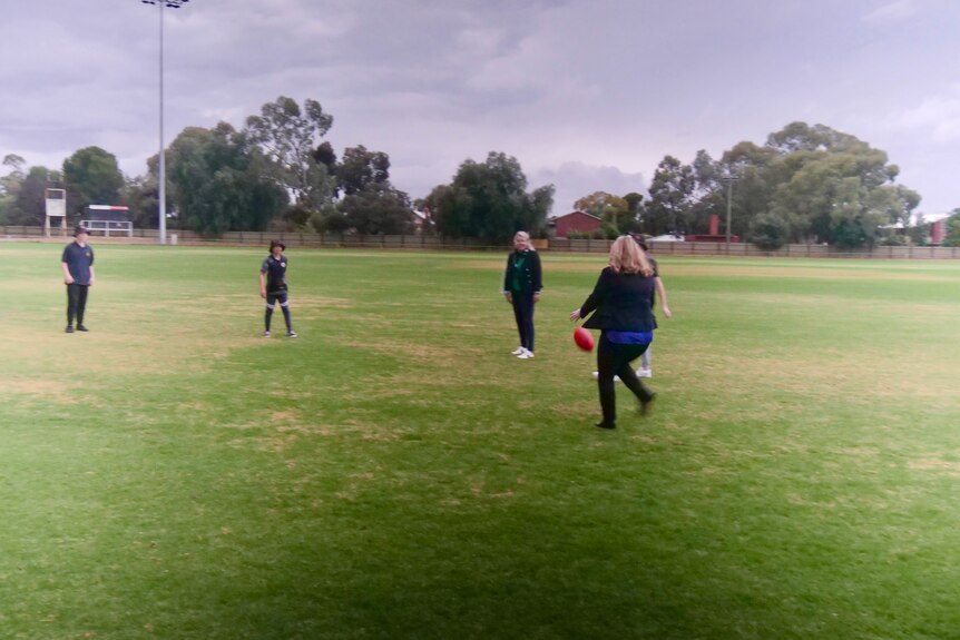 Woman kicking football on a green oval with school students. 