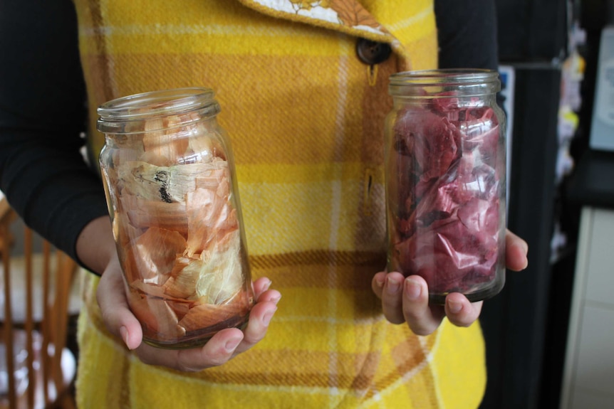 A person holds two jars in their hands, one full of red onion skins and the other full of white onion skins.