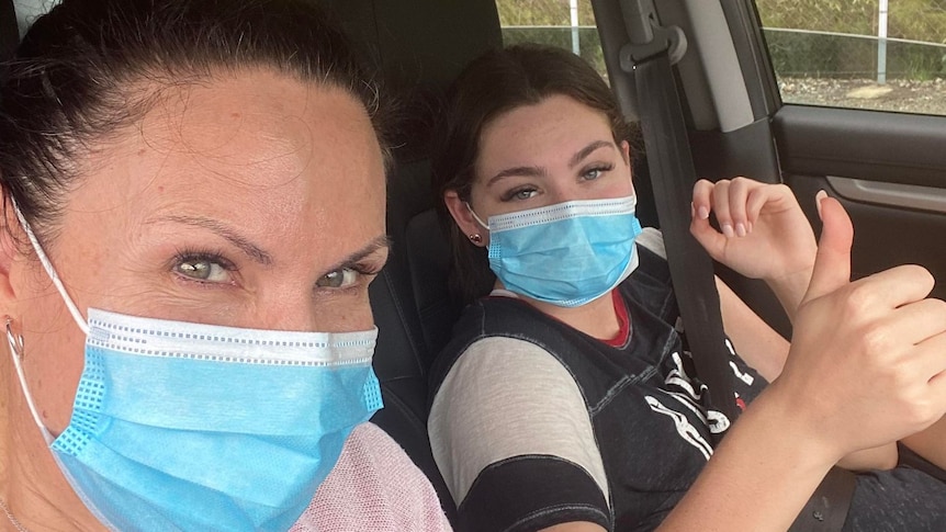 A masked woman and teen girl sitting in a car give thumbs up to the camera.