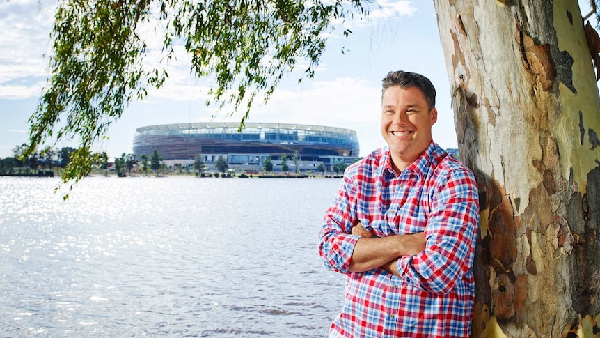 A man with short hair and a checked shirt, leaning against a gum tree, with water and a stadium in the background.