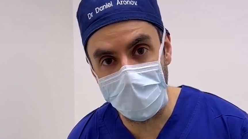 A man wearing a surgeon's scrubs, mask and a hate with his name embroidered looks directly into the camera.