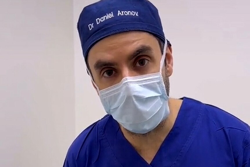 A man wearing a surgeon's scrubs, mask and a hat with his name embroidered looks directly into the camera.