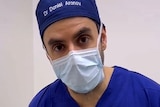 A man wearing a surgeon's scrubs, mask and a hate with his name embroidered looks directly into the camera.